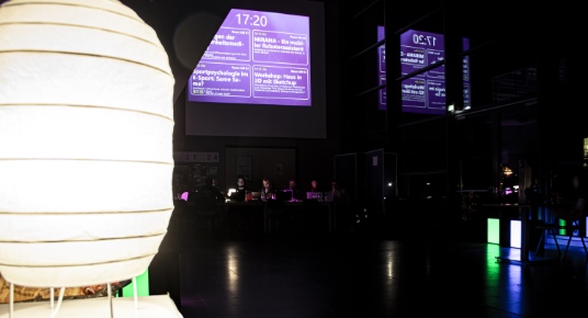 Foyer des Audimax im dunkeln. Links im Vordergrund eine Lampe. Im Hintergrund wird auf einem Beamer der Schedule angezeigt.